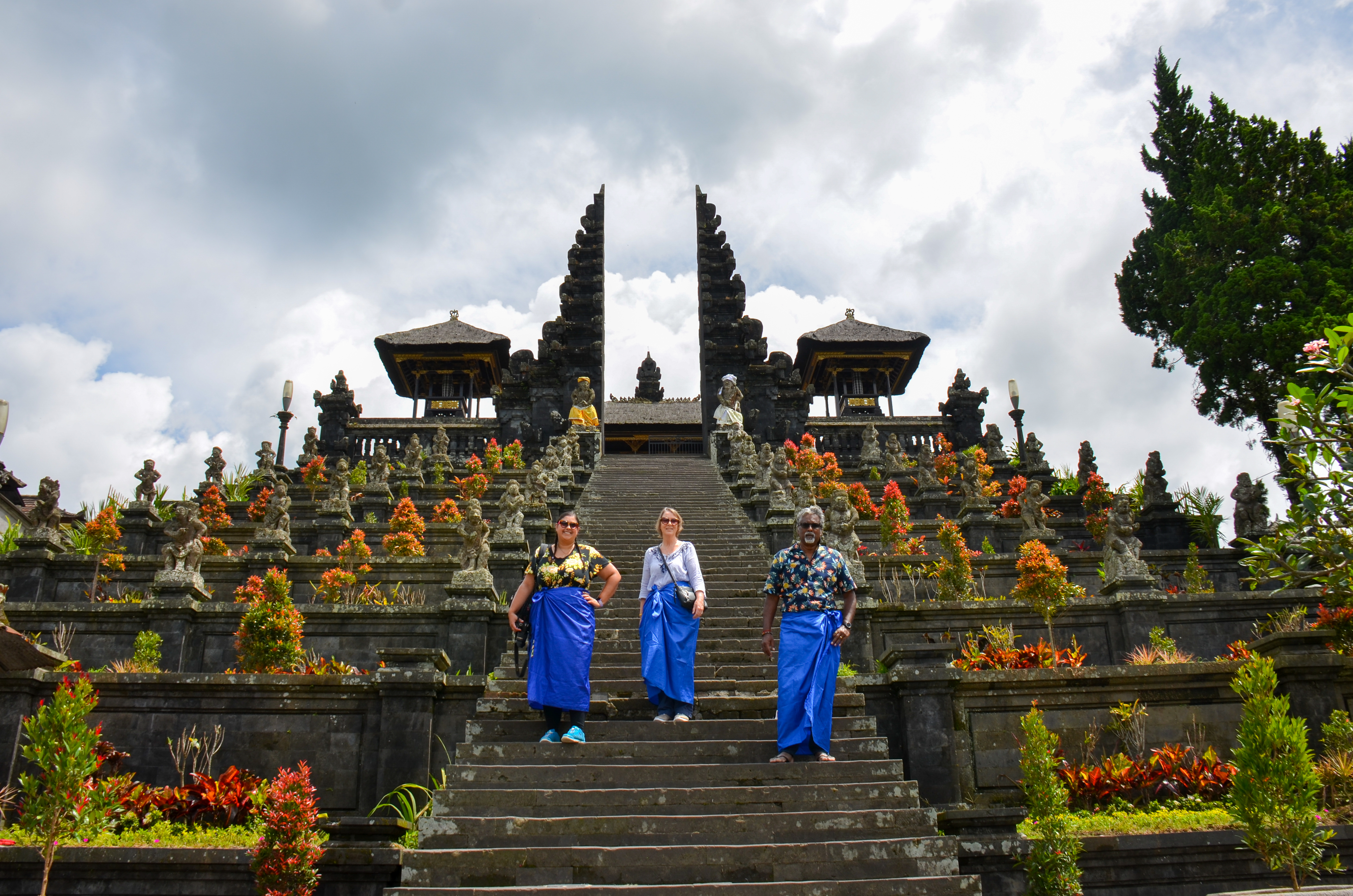 Three travelers standing on the steps of a Balinese temple, surrounded by lush greenery and colorful flowers, with traditional architecture visible in the background under a cloudy sky.