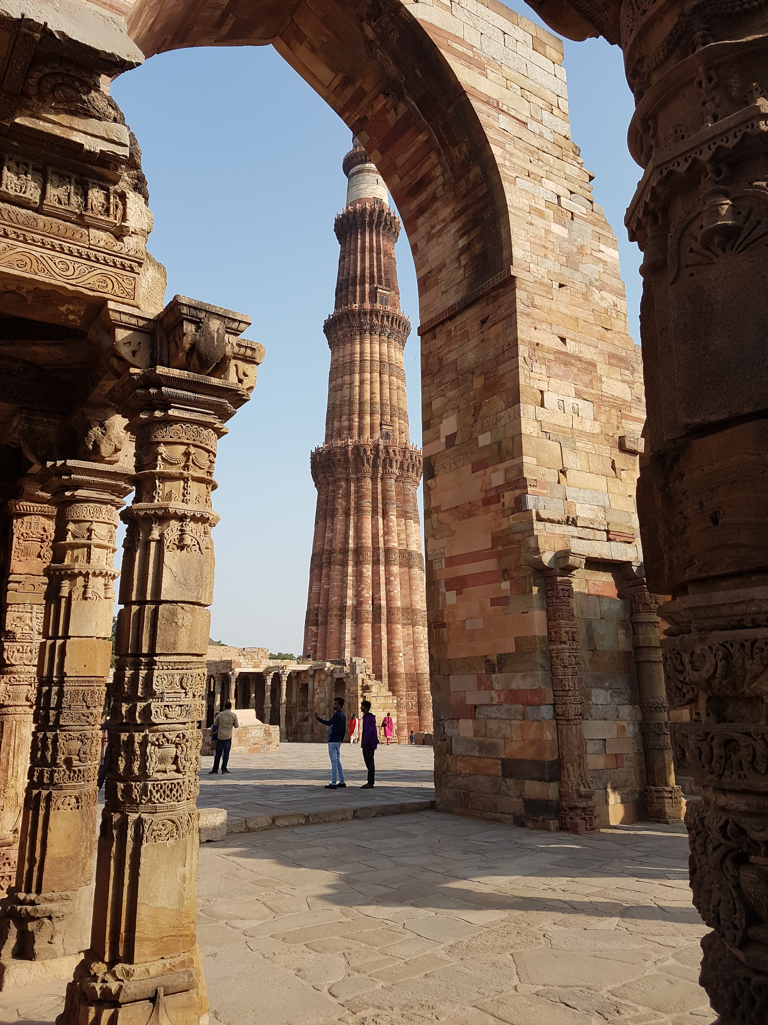 A view of the Qutub Minar, a tall red sandstone tower, framed by ancient architectural arches and ornate columns.
