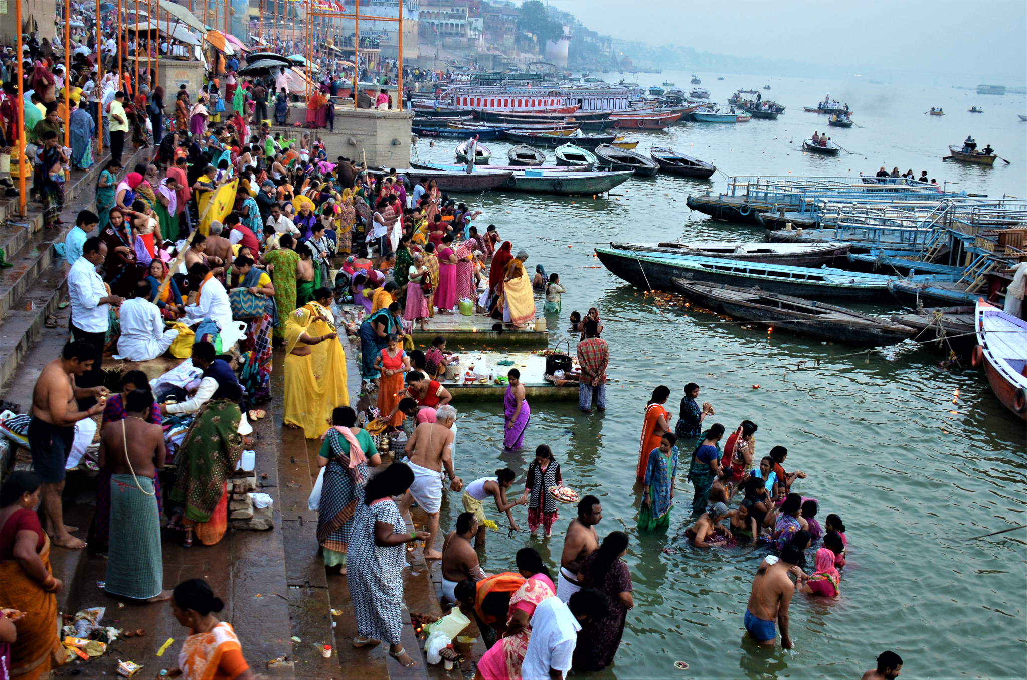 A vibrant scene at the Ghats of Varanasi, with people bathing and performing rituals in the Ganges River, surrounded by colorful clothing and boats.