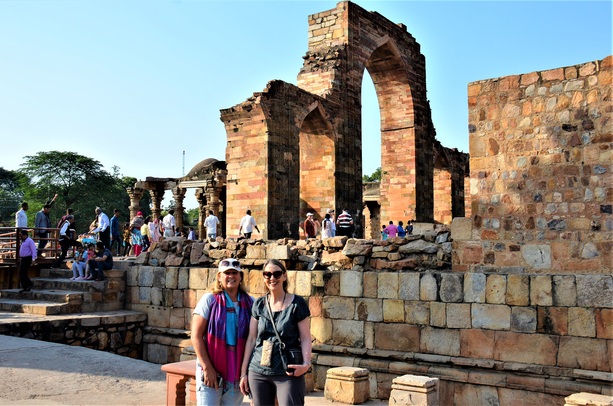 Two women posing in front of ancient ruins at a historical site in India, with visitors exploring the area.