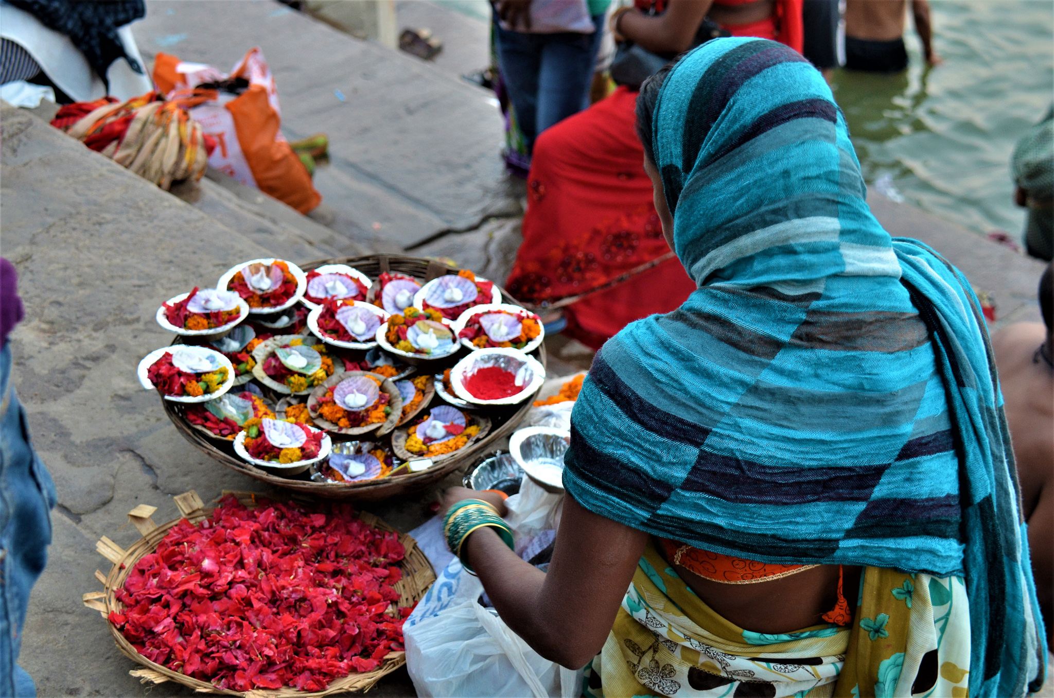 A woman wearing a blue scarf gathers flower offerings at the riverbank in Varanasi, surrounded by colorful bowls filled with flowers and decorative items.