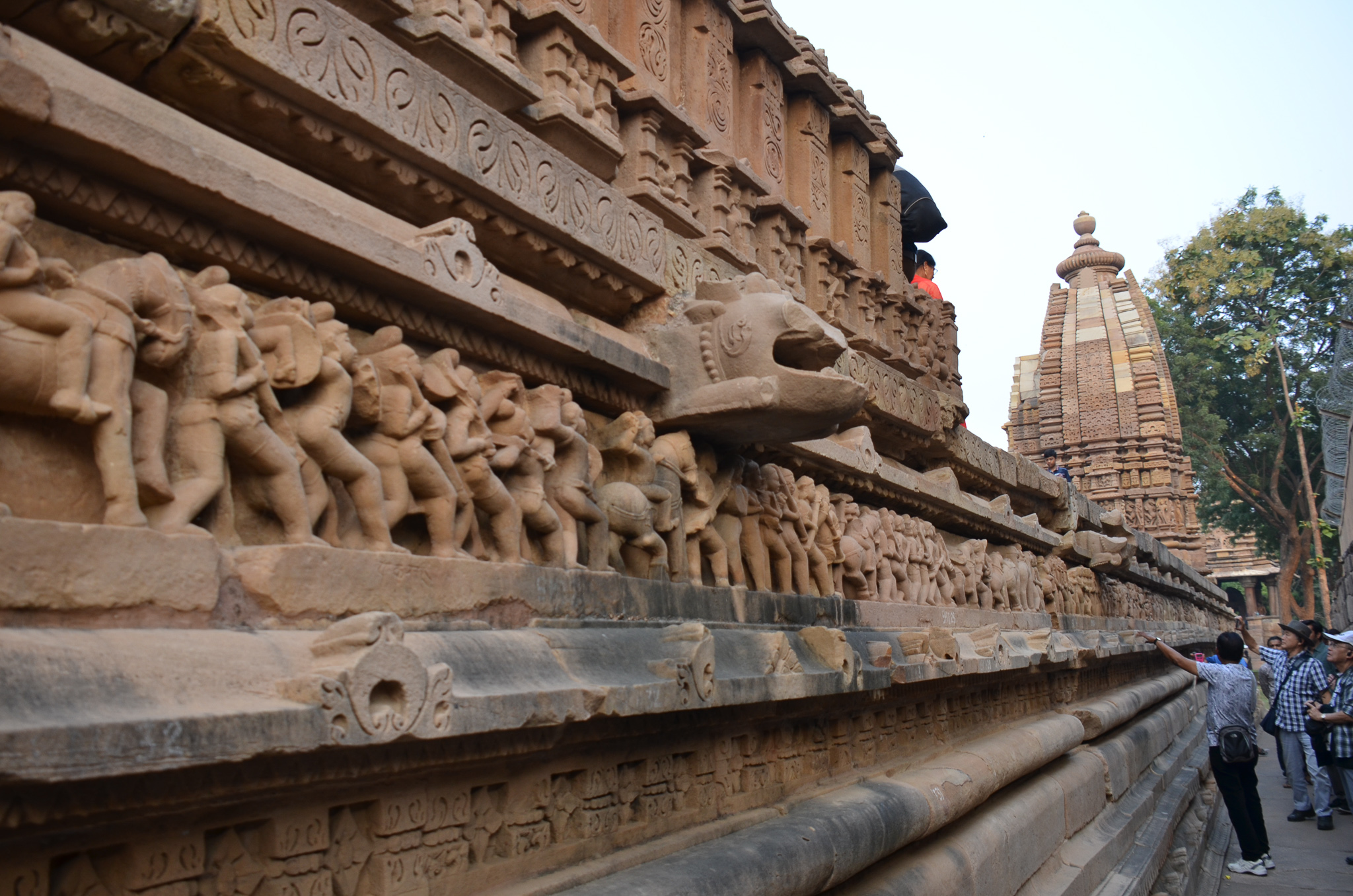 Close-up view of intricate carvings on the exterior wall of a temple in Khajuraho, India, depicting various figures and animals, with a temple spire in the background.
