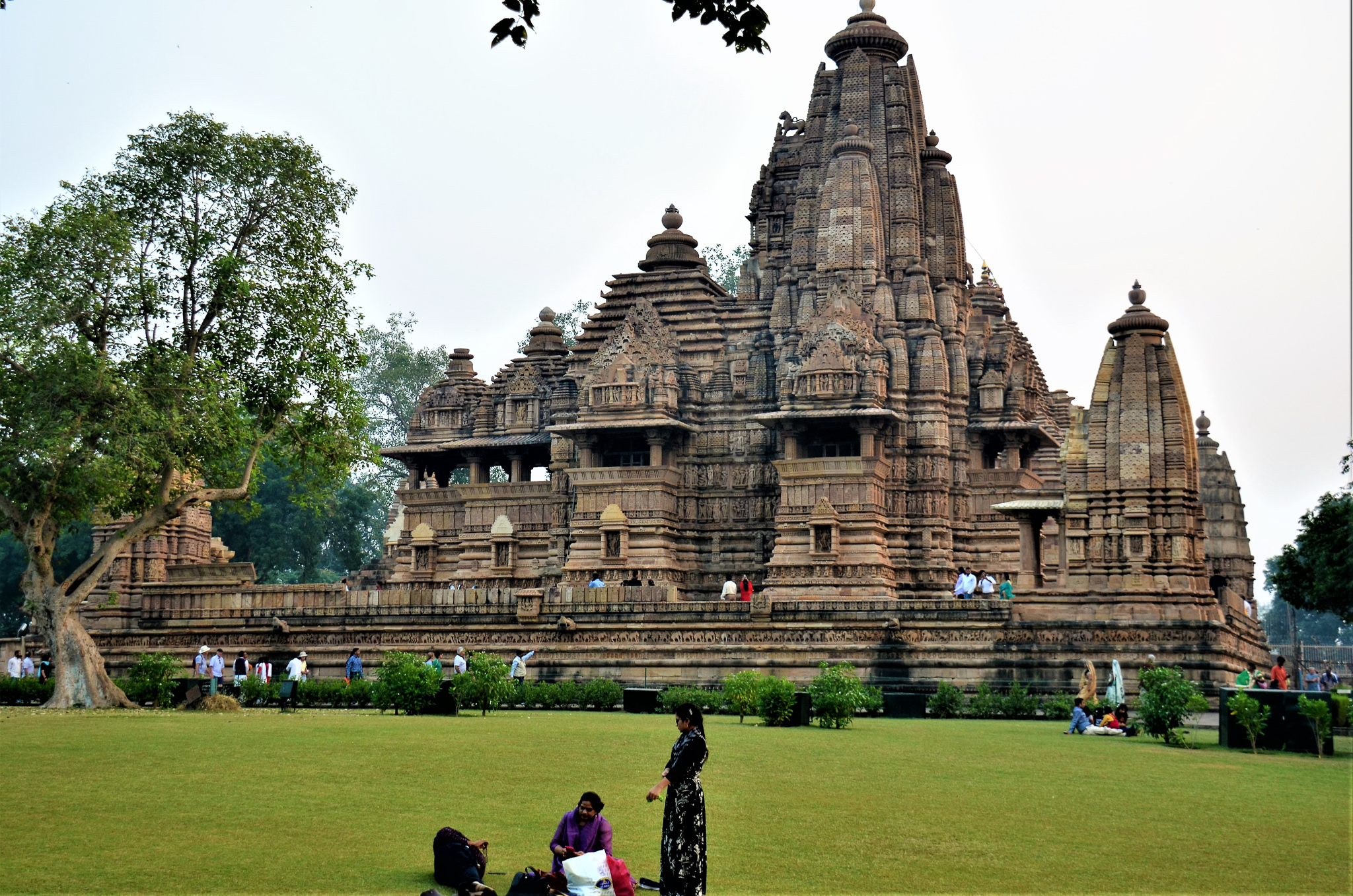 A view of a historic temple complex in Khajuraho, showcasing ornate architecture and intricate carvings, with visitors exploring the area on a green lawn.