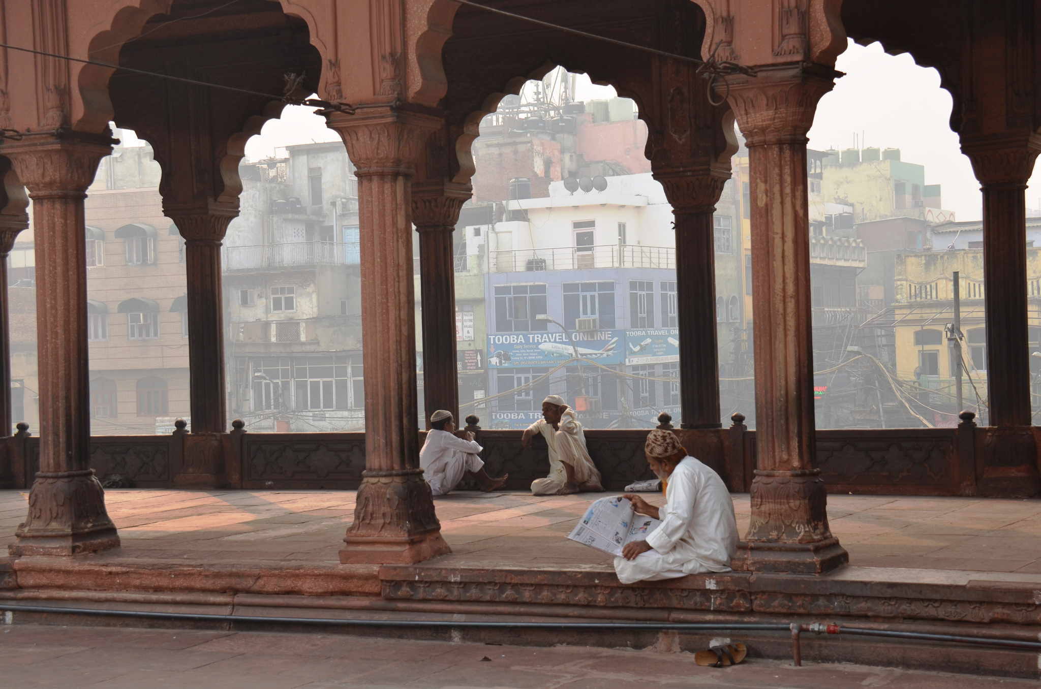 Three men seated in a traditional pavilion in Old Delhi, surrounded by ornate pillars, with urban buildings visible in the background.