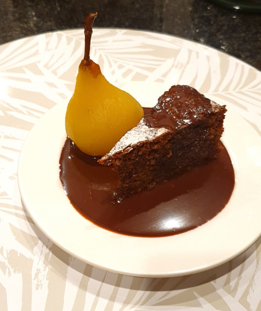 A rich, decadent chocolate torte with a slightly cracked top, dusted with cocoa powder, served on a simple plate alongside a drizzle of chocolate sauce and elegant white wine–poached pears. The dense, nutty interior of the torte contrasts beautifully with the glossy pear, creating a comforting yet refined dessert perfect for special occasions.