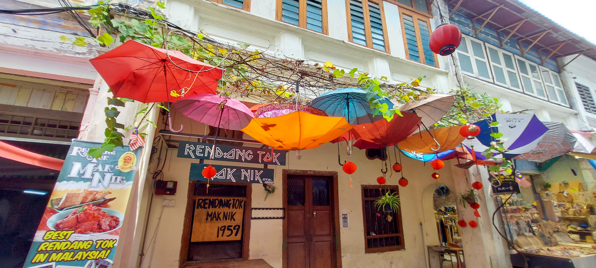 Colorful umbrellas hanging above a restaurant entrance with a sign reading 'Rendang Tok Mak Nik' in a historic street in Ipoh, Malaysia.