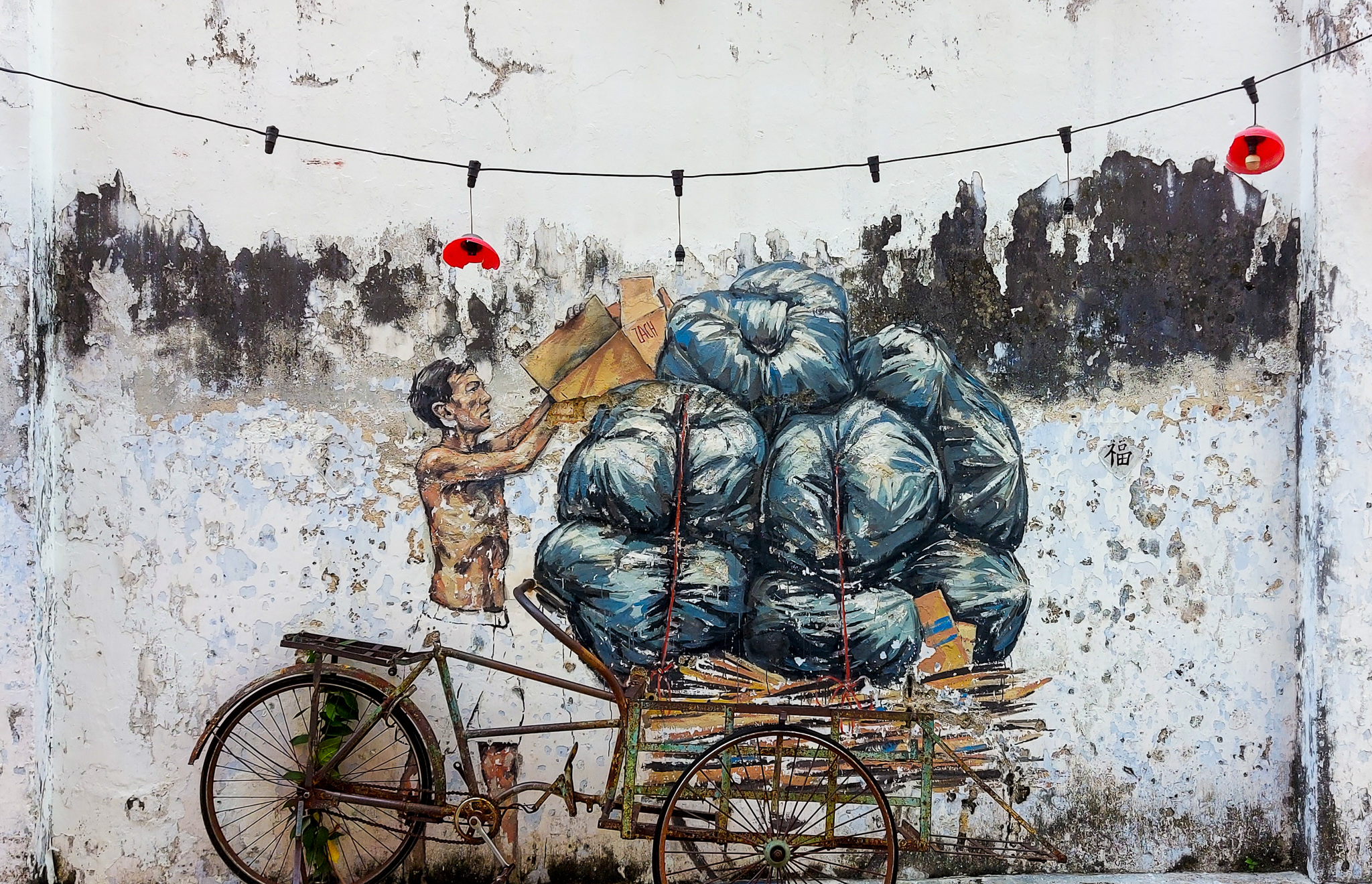 A mural depicting a man loading garbage bags onto a cart against a weathered wall, with decorative red lanterns hanging above.