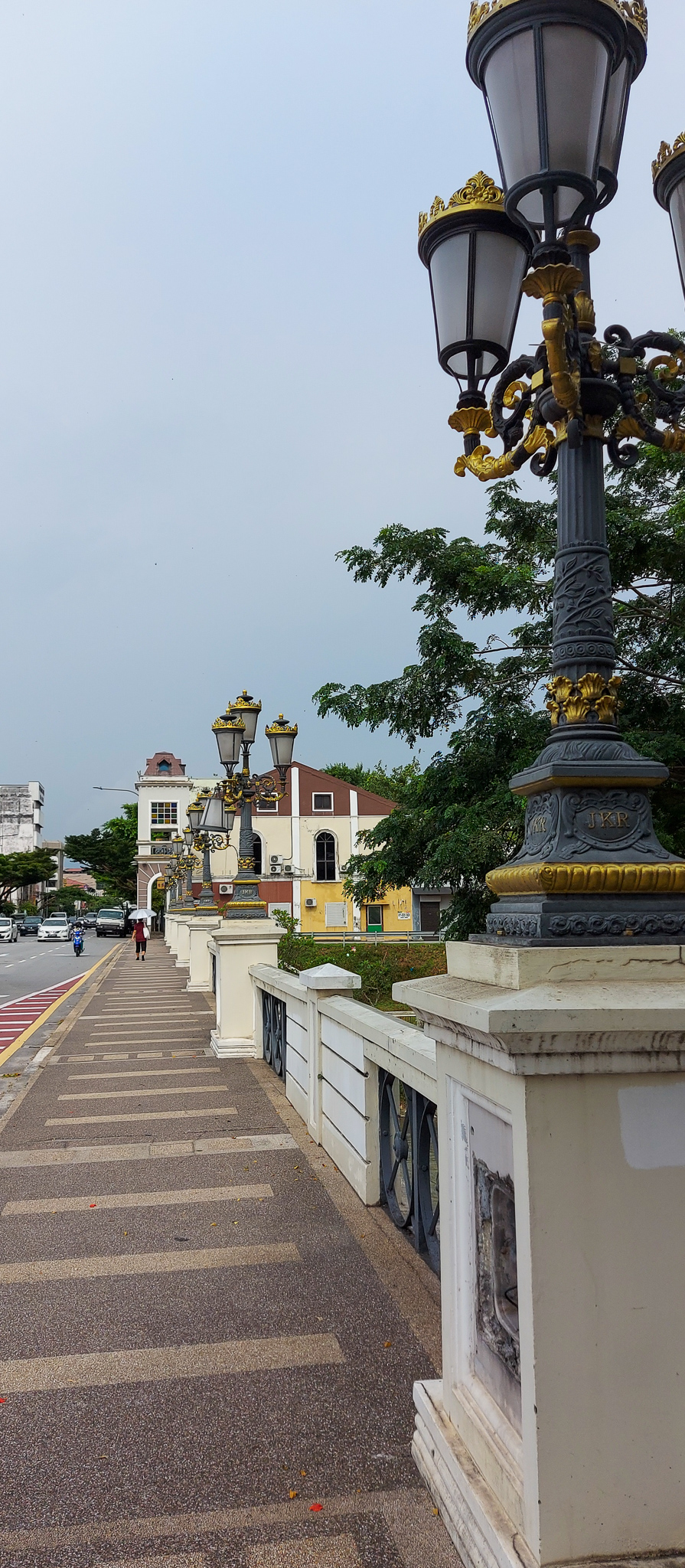 A walkway lined with decorative street lamps and a white railing, leading alongside a road in Ipoh, Malaysia. The background features historical buildings and lush green trees under a cloudy sky.