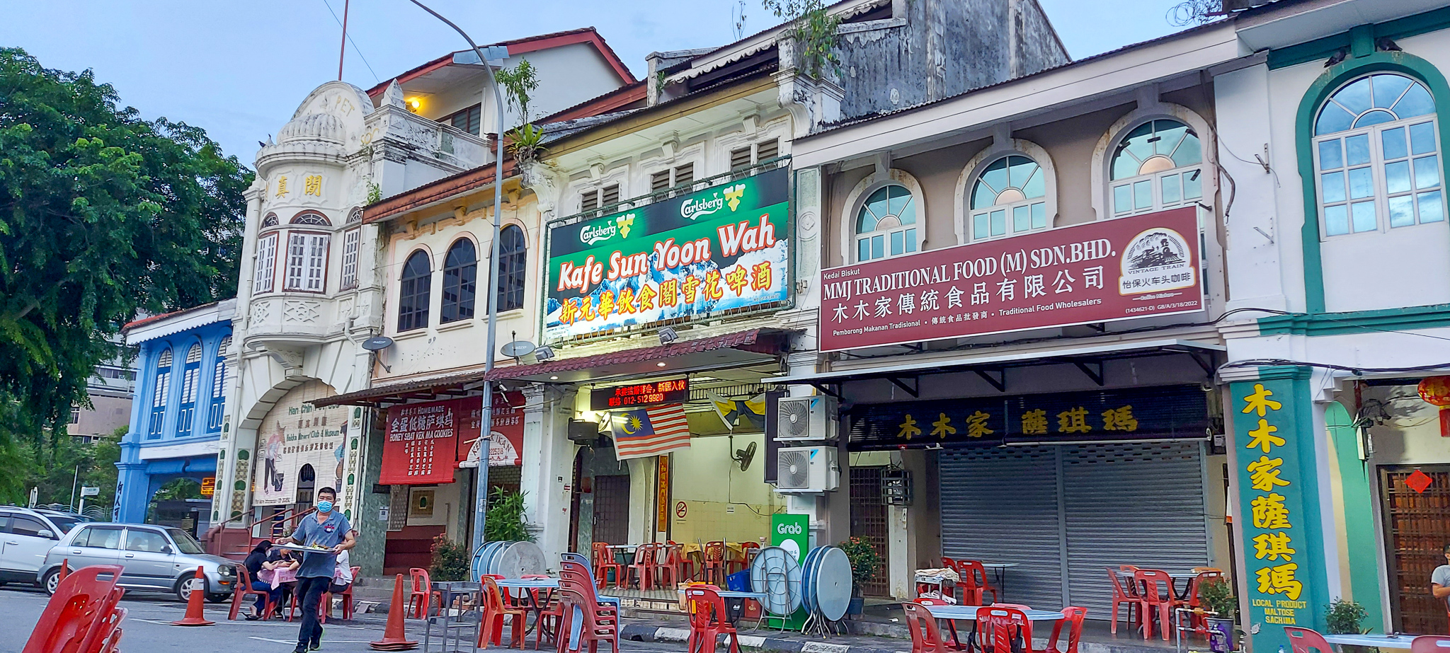 A street view of historic buildings in Ipoh, Malaysia, featuring colorful shop signs and outdoor dining tables with a man carrying a tray.