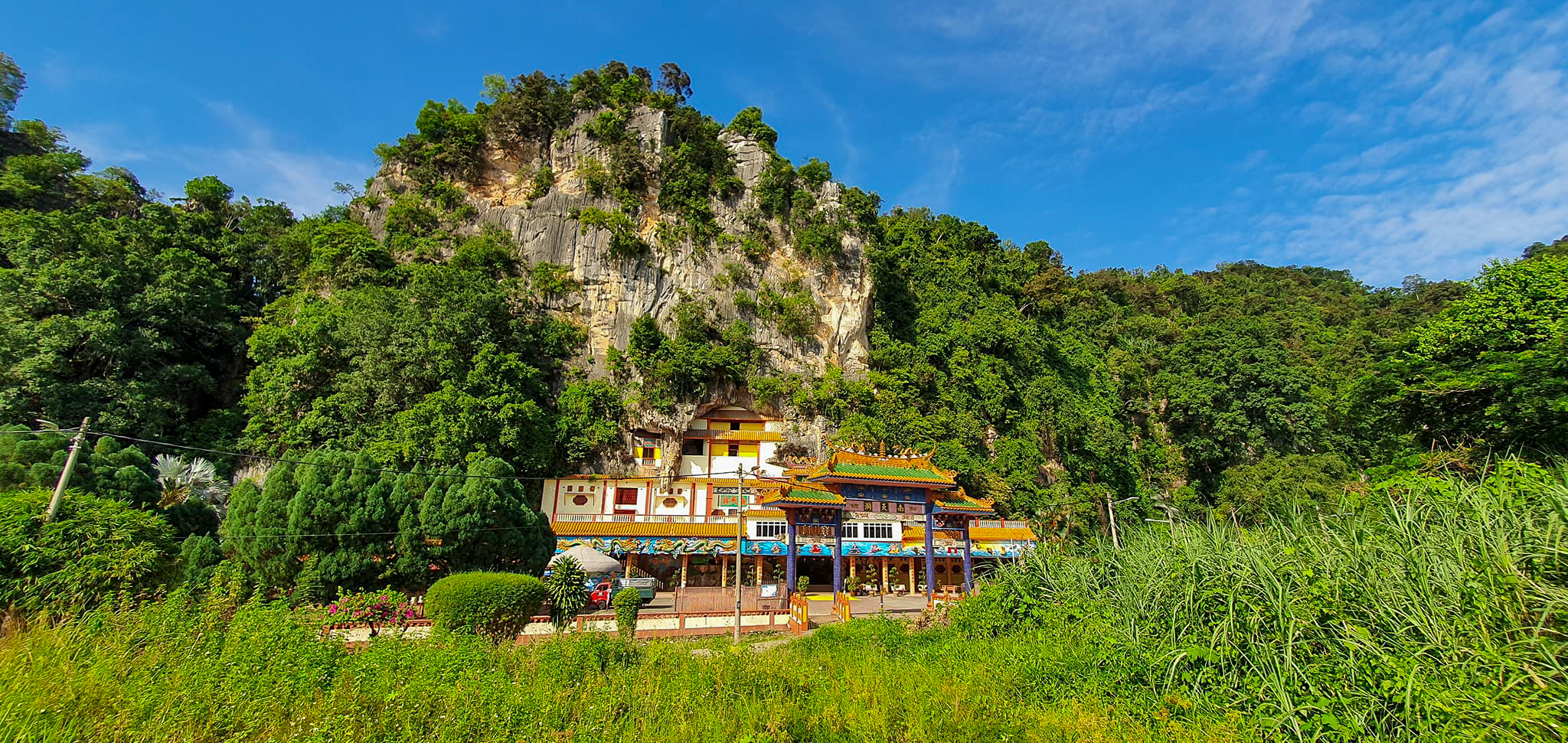 A scenic view of a colorful temple built at the base of a lush, rocky hill under a bright blue sky.