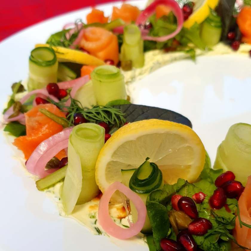 A smoked salmon wreath arranged on a serving platter, with layers of smoked salmon, creamy dollops of cream cheese, sprigs of fresh dill, crisp cucumber slices, and bright pickled onions. A colourful, crowd-pleasing starter that’s always a hit at family gatherings, holidays, or any special occasion.