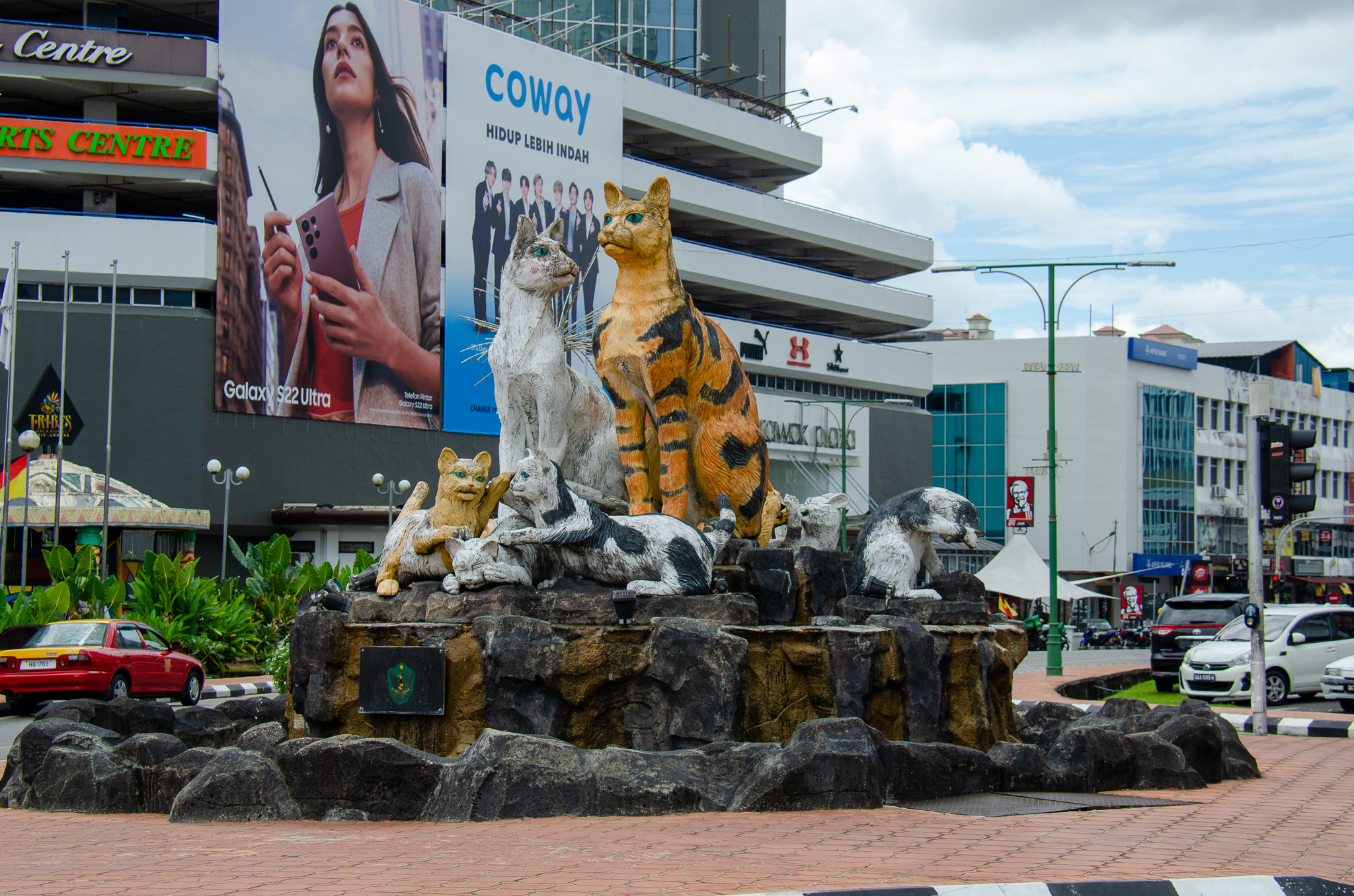 Statue of various cats, including a tiger and domestic cats, displayed on a rock formation in Kuching, Malaysia, with urban buildings and advertisements in the background.