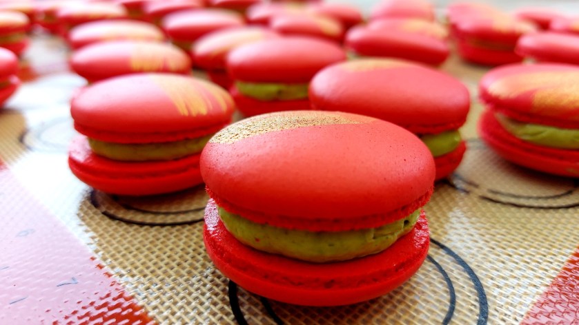Vibrant red macarons with a slight gold dusting and a green macha tea buttercream filling, still on the baking mat.