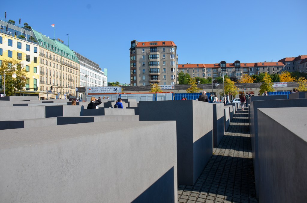 A view of the Holocaust Memorial in Berlin, featuring its grey concrete slabs arranged in a grid pattern, with people walking and exploring the site under a clear blue sky.