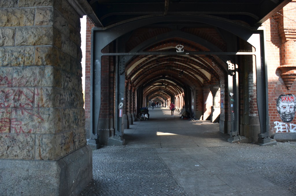 An arched corridor featuring brick and stone pillars, with graffiti on the walls, leading to a distant view of pedestrians.