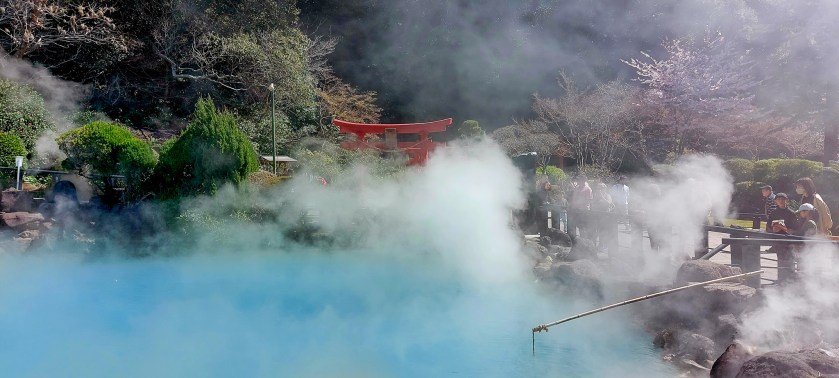 A scenic view of a hot spring in Japan with steam rising from the colorful water, surrounded by lush greenery and a traditional red torii gate in the background.