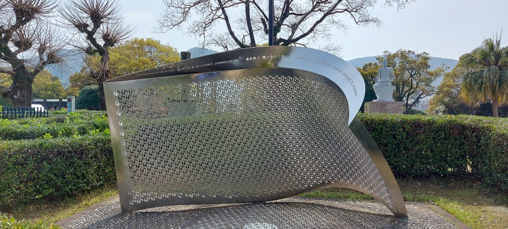 A modern memorial sculpture made of stainless steel, set in a landscaped park with trees and greenery in the background.