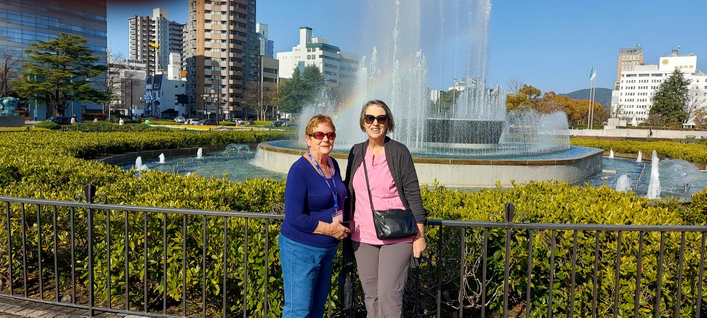 Two women stand in front of a large fountain surrounded by greenery and buildings, smiling for the camera on a sunny day.