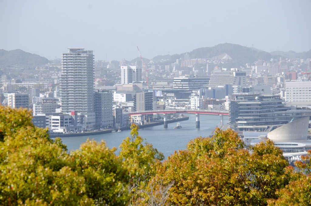 A panoramic view of a city skyline featuring modern buildings, a bridge crossing a river, and distant hills under a clear sky.