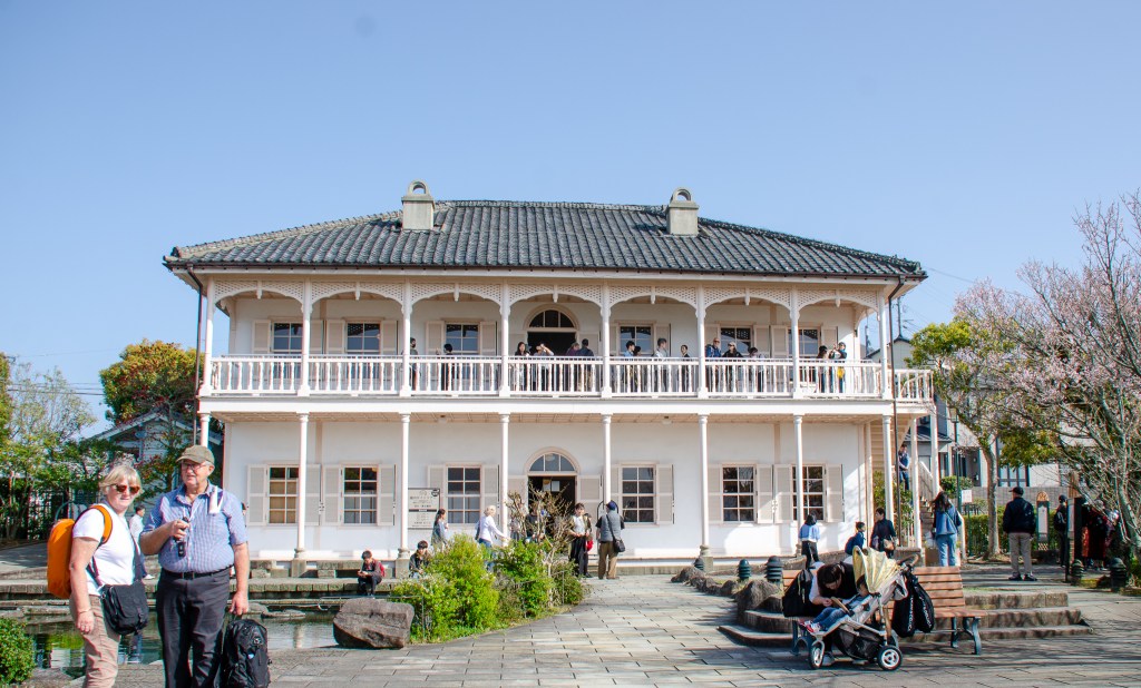 A historic white building with a second-floor balcony, surrounded by greenery and cherry blossoms. Two tourists stand in the foreground smiling at the camera.
