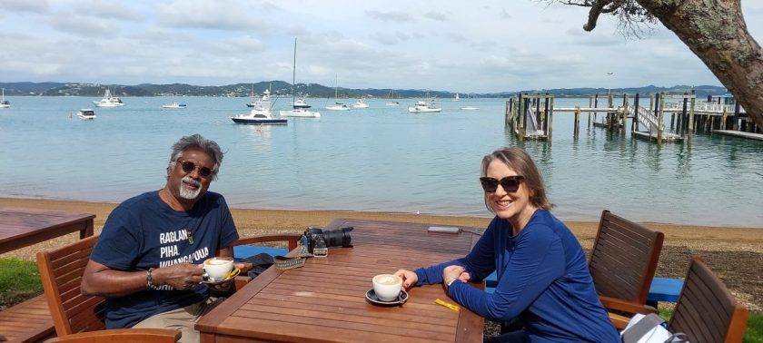 Two people sitting at an outdoor table near the beach, having cups of coffee, with calm water and boats in the background.