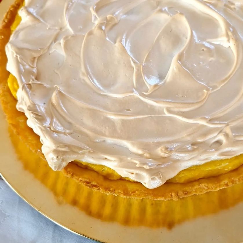 A pineapple tart topped with golden, toasted meringue swirls, served on a decorative plate with a flaky pastry crust and glossy fruit filling.
