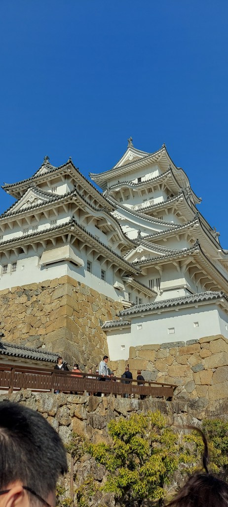 A view of Himeji Castle against a clear blue sky, showcasing its elegant architecture and traditional Japanese design, with visitors gathered in front.