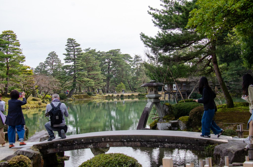 Visitors capturing the serene beauty of a traditional Japanese garden featuring a pond, stone lanterns, and lush greenery.