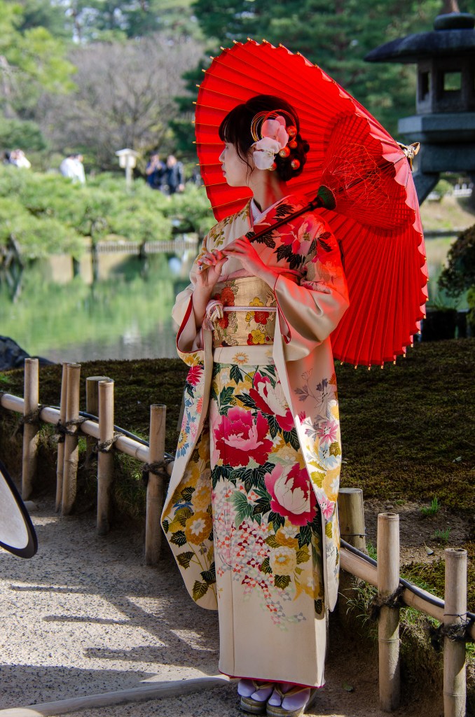 A woman in a colorful kimono stands near a serene pond with a red parasol, surrounded by lush greenery.