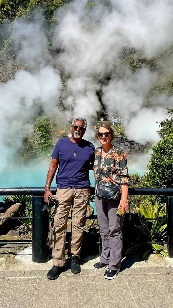 A couple poses for a photo in front of steaming hot springs, surrounded by lush greenery.