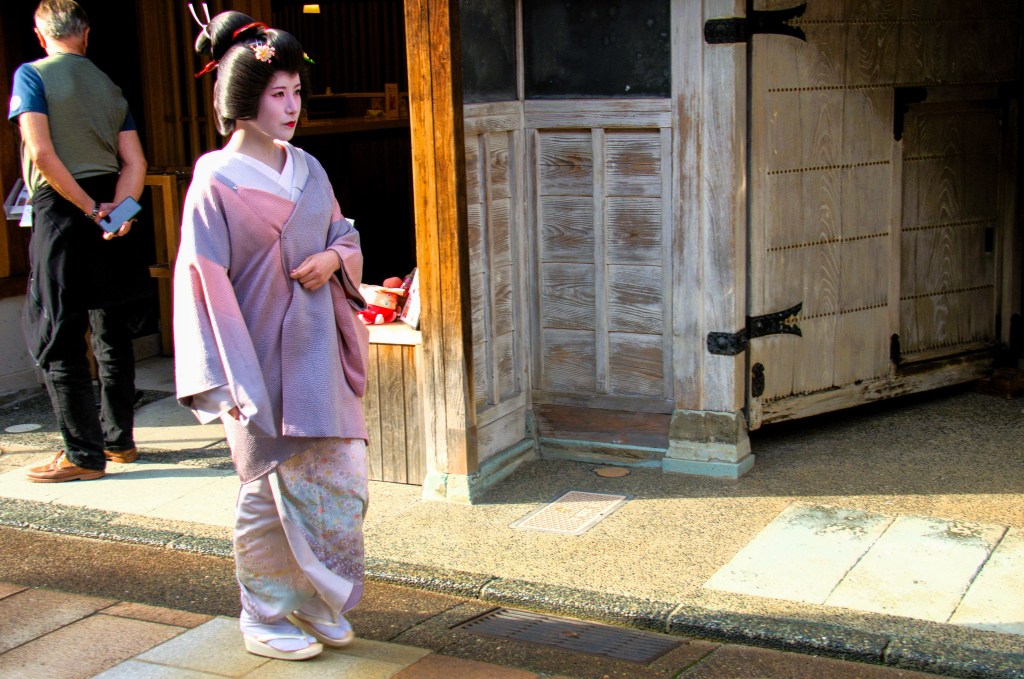 A young woman wearing a traditional Japanese kimono stands gracefully on a street, near a wooden building, contributing to the cultural ambiance of the area.