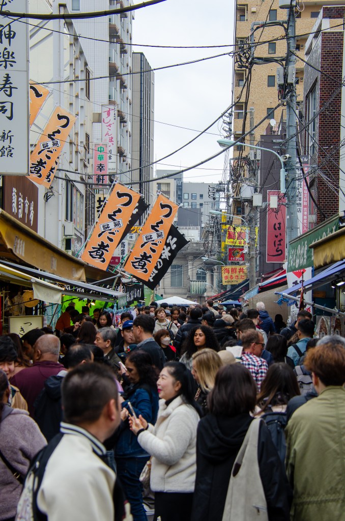A bustling street market in Japan filled with a diverse crowd of people exploring food stalls and shops, with colorful banners hanging overhead.