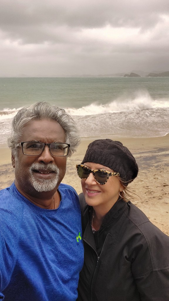 A couple posing for a selfie at a beach on a cloudy day, with rough sea waves in the background.