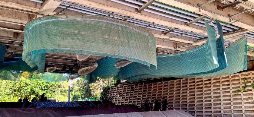 Art installation with flowing translucent elements under a bridge, surrounded by greenery and people observing.