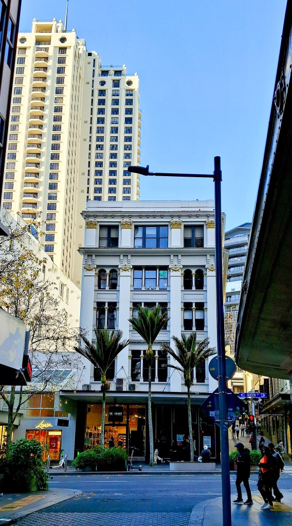A view of a historical building with palm trees in front, contrasting against a modern high-rise in an urban street setting.