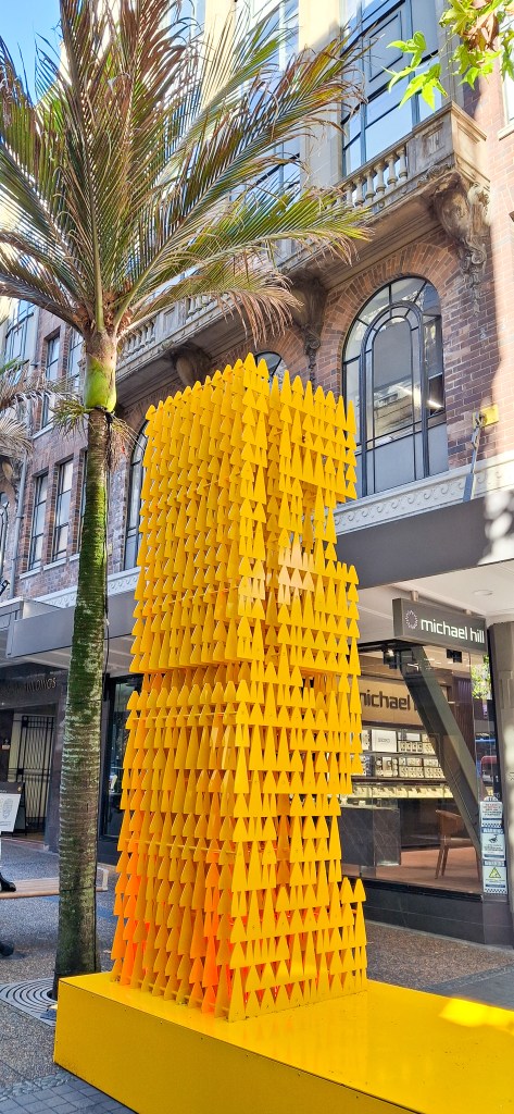 A colorful yellow art installation made of triangular shapes displayed outdoors in a city setting, with a palm tree and buildings in the background.