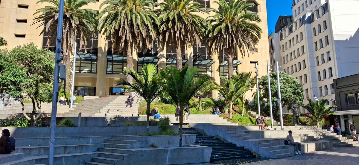 A sunny urban scene featuring a staircase surrounded by palm trees, with people sitting and relaxing on the steps in front of a modern building.