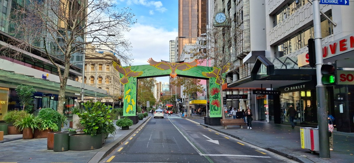 A vibrant street view of Queen Street featuring Matariki decorations, showcasing an ornate arch with indigenous designs and artwork, surrounded by shops and greenery.