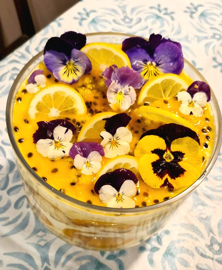 A beautifully layered lemon and peach trifle topped with edible flowers and lemon slices, served in a glass bowl.