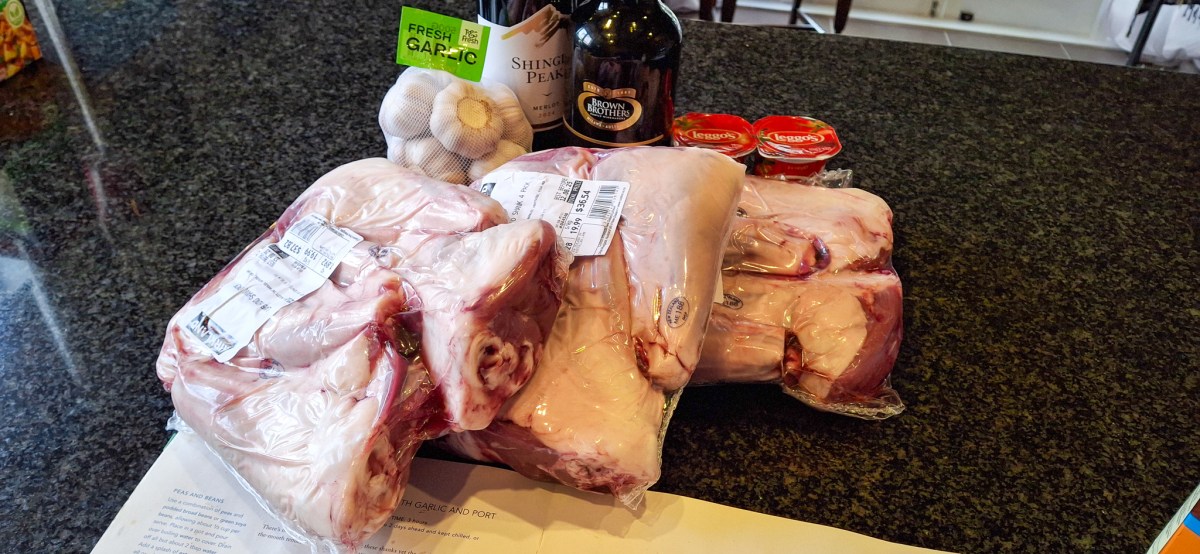 Ingredients for Slow-Baked Lamb Shanks including packaged lamb shanks, garlic, red wine, and sauce ingredients on a kitchen counter.