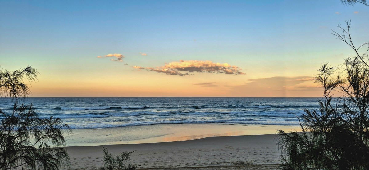 A tranquil beach scene at sunset, with calm waves lapping at the shore and a colorful sky above, framed by coastal vegetation.
