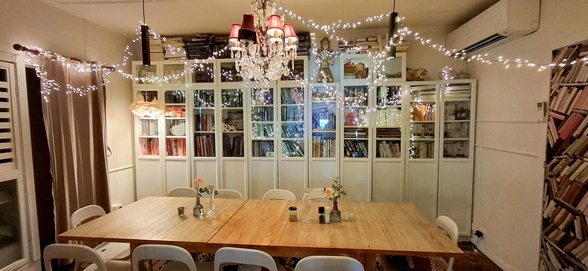 Cozy dining area with a large wooden table, surrounded by white chairs, and decorated with fairy lights and a chandelier, creating a warm ambiance.