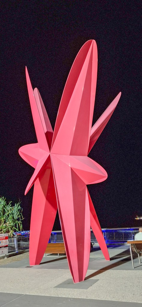 A large, modern pink sculpture resembling a star, set against a dark night background with city lights in the distance.