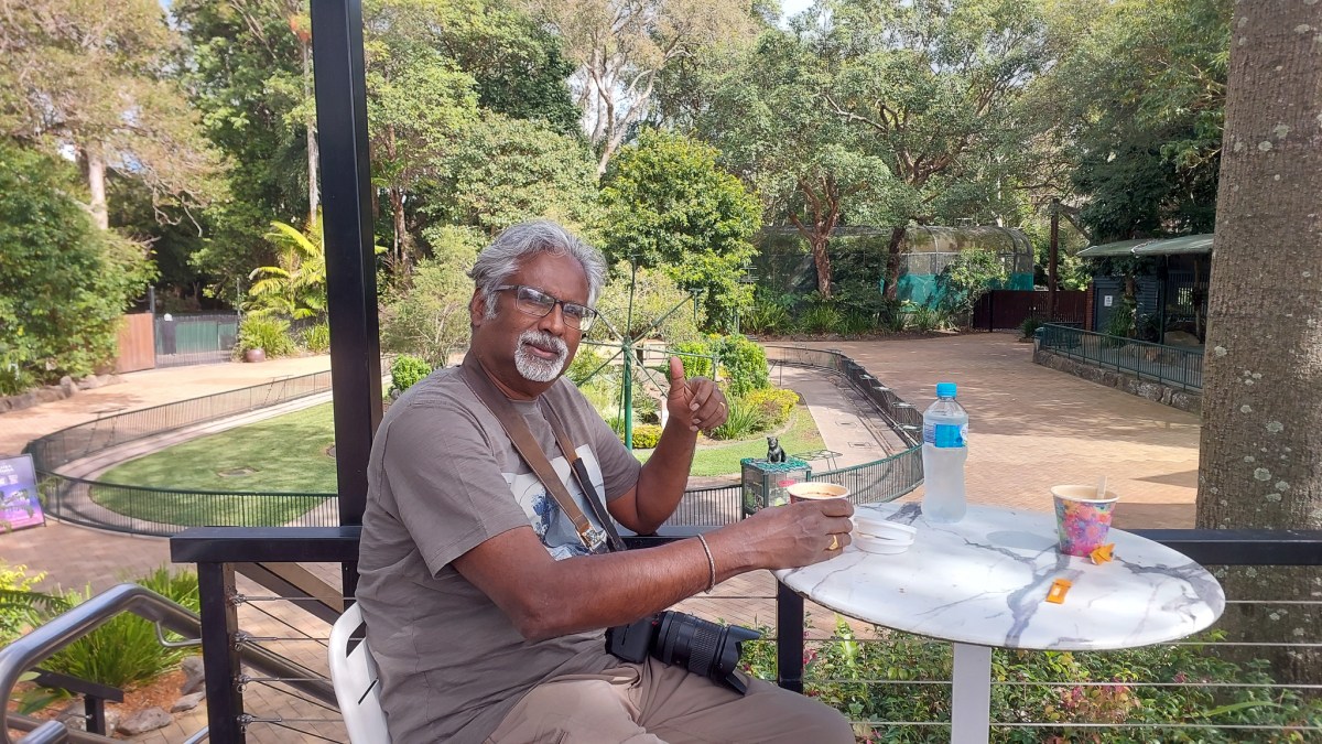 A man sitting at a table with a cup, giving a thumbs up gesture, surrounded by greenery at a wildlife sanctuary.