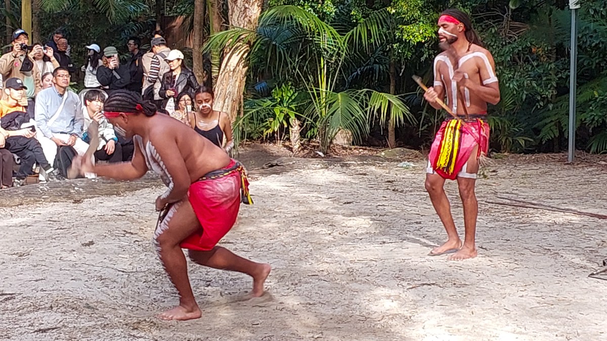 Two performers in traditional dress engaging in a cultural dance in front of an audience amidst lush greenery.