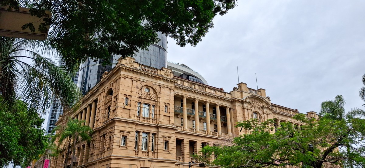 Historic building in Brisbane, Australia, featuring ornate architecture, surrounded by greenery and modern skyscrapers under a cloudy sky.
