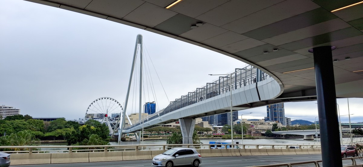 A view of a modern bridge spanning over a river, with a large observation wheel visible in the background and cityscape elements around.