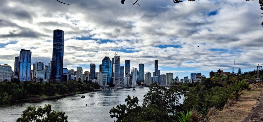 A panoramic view of Brisbane's skyline along the river, featuring tall skyscrapers and cloudy skies.