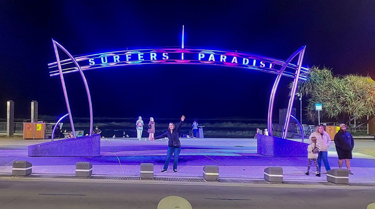 Night view of the Surfers Paradise entrance sign, illuminated with colorful lights. A person poses in the foreground while others are seen enjoying the beach atmosphere in the background.
