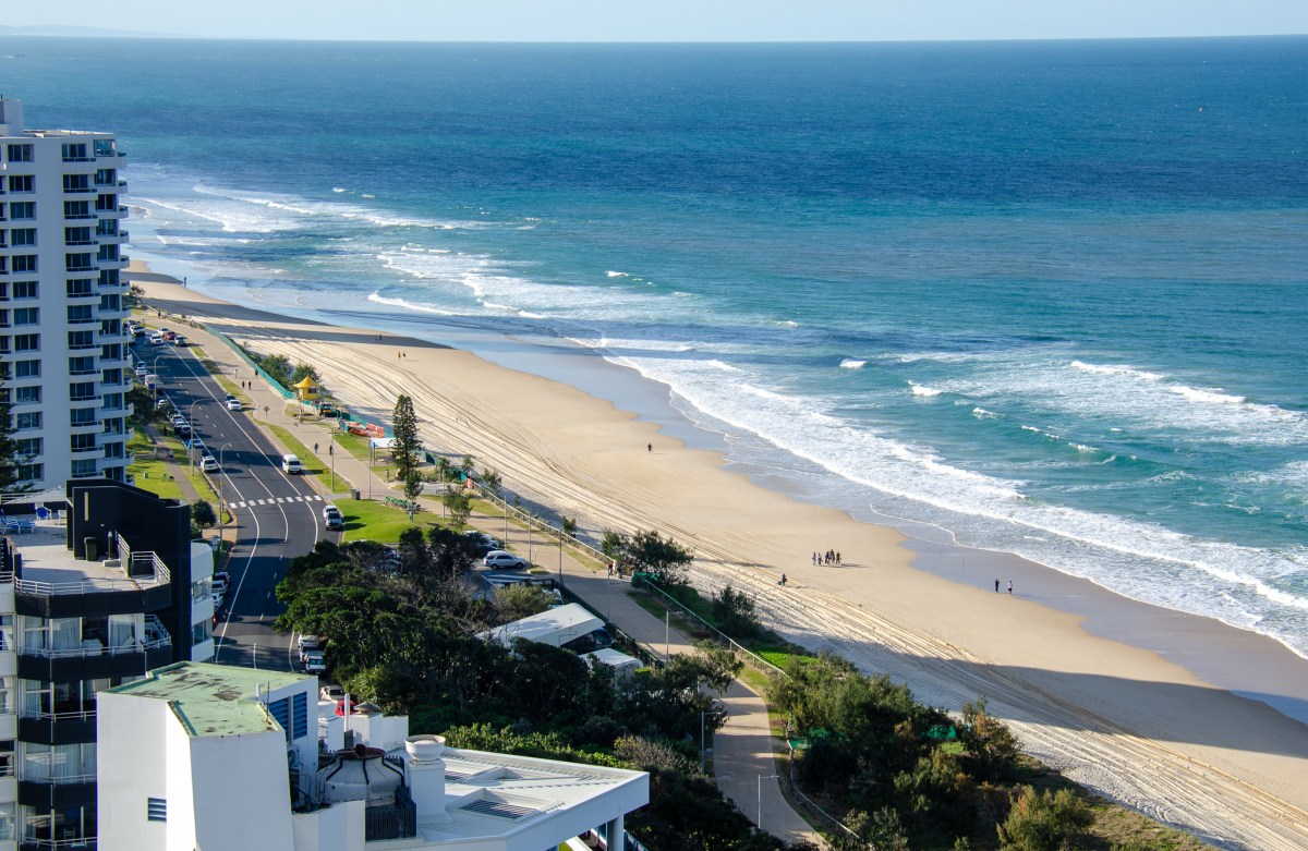 View of the sandy beach and ocean at Gold Coast, Australia, with people walking along the shoreline and modern buildings nearby.